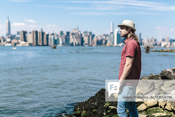 USA New York City Mann mit Hut am Wasser mit Skyline von Manhattan im Hintergrund