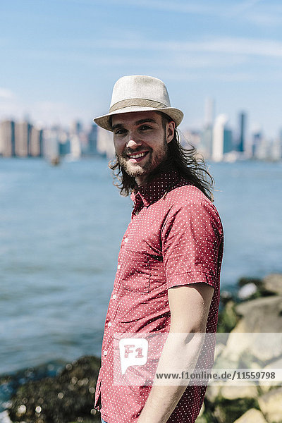 USA  New York City  lächelnder Mann mit Hut am Wasser mit Manhattan-Skyline im Hintergrund