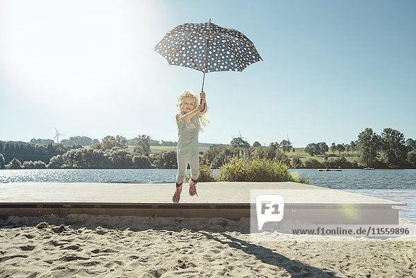 Mädchen mit Regenschirm  das an einem sonnigen Tag von einem Steg springt.