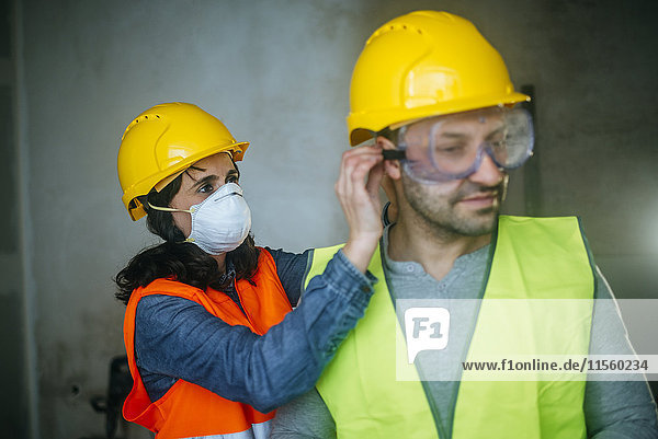 Frau hilft Mann beim Aufsetzen einer Schutzbrille auf einer Baustelle