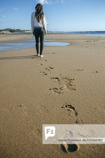 Rückansicht einer jungen Frau  die am Strand spazieren geht und ihre Fußspuren im Sand hinterlässt.