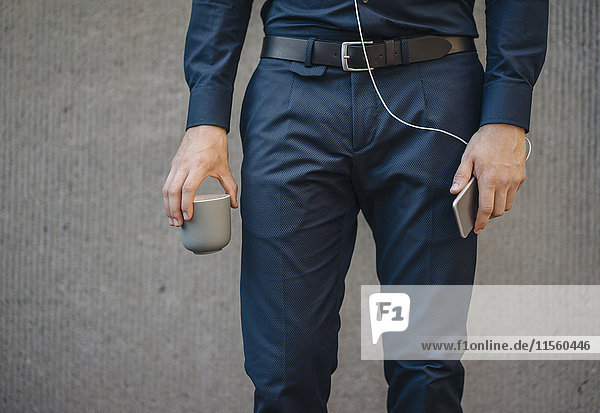 Geschäftsmann hält Smartphone mit angeschlossenen Kopfhörern und Tasse Kaffee
