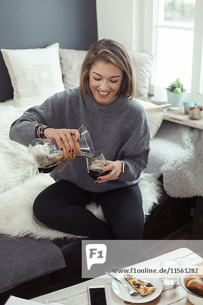 Lächelnde blonde Frau sitzt auf der Couch und gießt Kaffee in ein Glas.