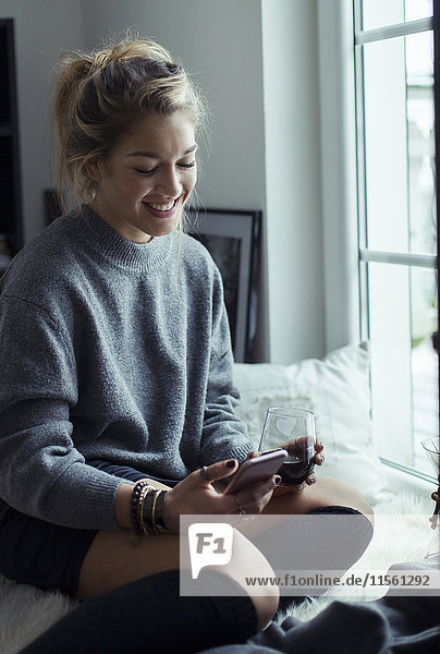 Lächelnde junge Frau mit Tasse Kaffee entspannt auf Schaffell zu Hause beim Blick auf Handy