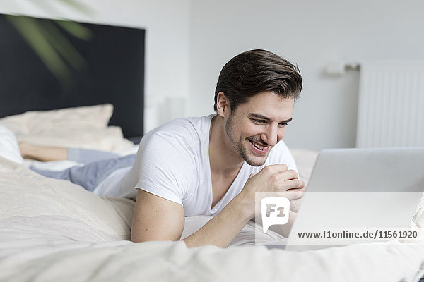 Smiling man lying on bed using laptop