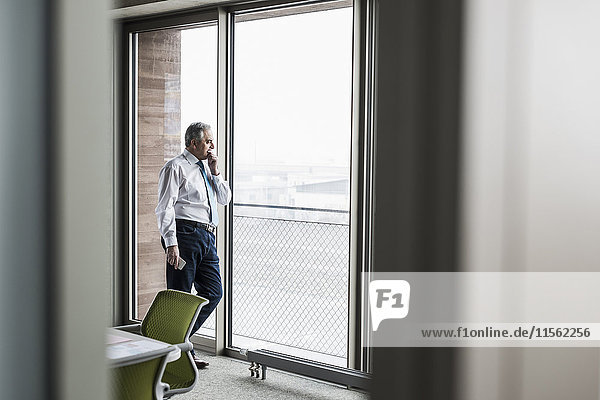Senior Manager im Büro stehend am Fenster mit Smartphone