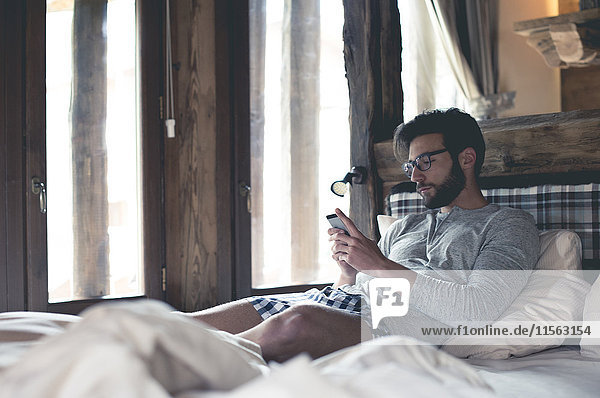 Junger Mann mit Smartphone zu Hause im Bett liegend