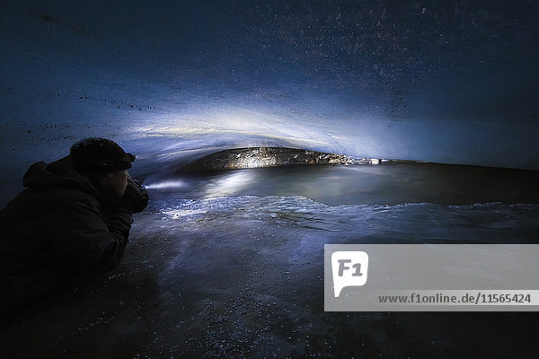 'A man in a cramped cave inside Fels Glacier (commonly misspelled as ''Eel'' Glacier) shines a light on a stream flowing beneath the glacier in the winter; Alaska  United States of America'