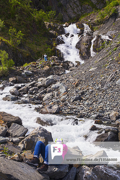 'A woman taking a break from sea kayaking in Kenai Fjords National Park relaxes by a rushing stream while her partner explores a waterfall; Alaska  United States of America'