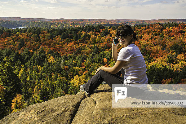 Frau spricht mit einem Smartphone  während sie auf einer Klippe im Algonquin Park sitzt; Ontario  Kanada'.