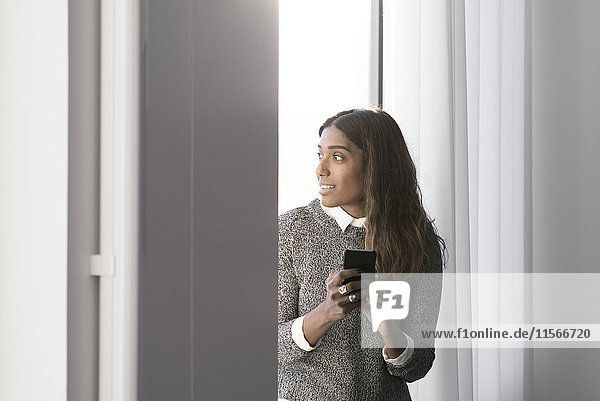 Frau hält Smartphone und schaut durch das Fenster