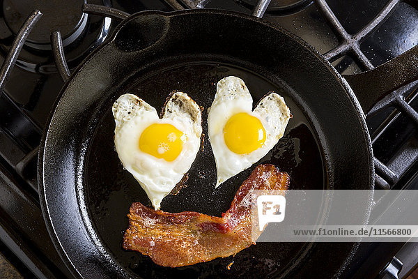 'Close up of heart shaped sunny side up eggs and a piece of bacon in a cast iron frying pan; Calgary  Alberta  Canada'