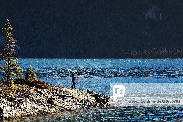 'Female hiker standing on rocky banks of a mountain lake with evergreen covered mountain in the background; Banff  Alberta  Canada'