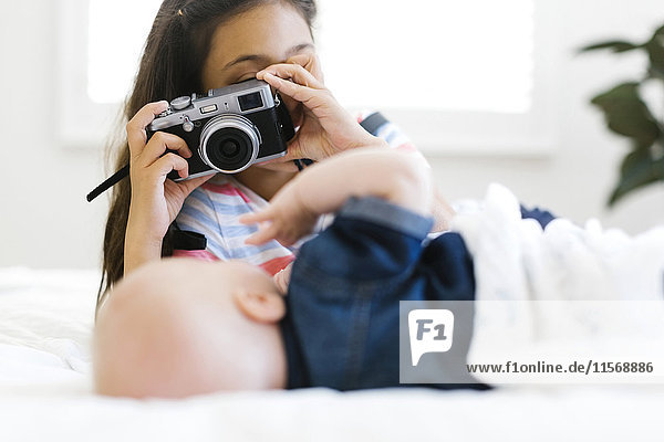 Girl (10-11) taking photo of her small brother (12-17 months) lying on bed