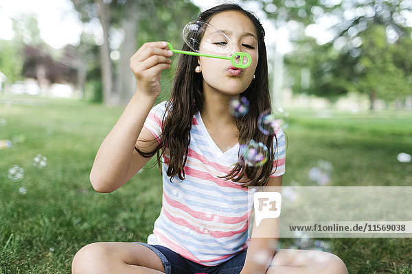 Porträt eines Mädchens (10-11)  das auf einer Wiese sitzt und Seifenblasen pustet