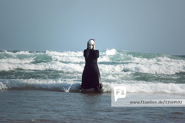 Frau in schwarzem Kleid mit Maske  stehend in der Brandung im Meer