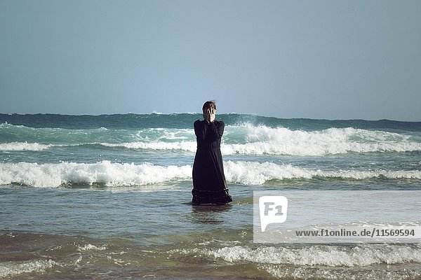 Frau in schwarzem Kleid mit Maske  stehend in der Brandung im Meer