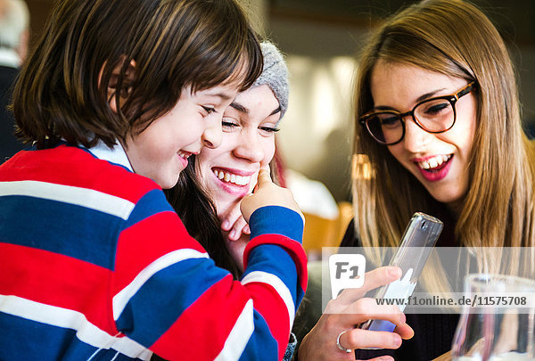 Zwei junge Frauen und ein Junge schauen und lachen über ein Smartphone im Cafe