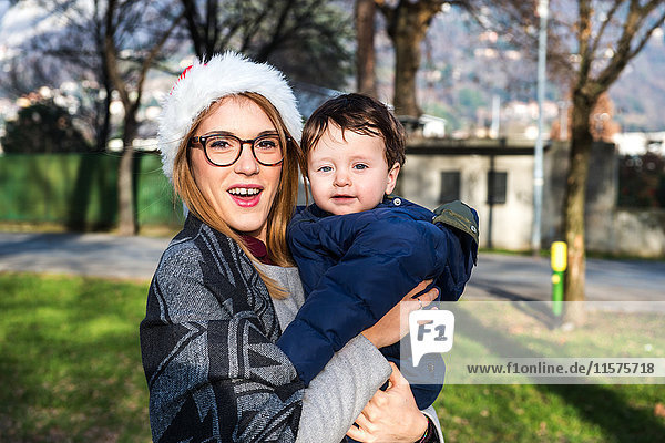 Porträt einer jungen Frau mit Weihnachtsmannhut  die einen kleinen Jungen im Park trägt