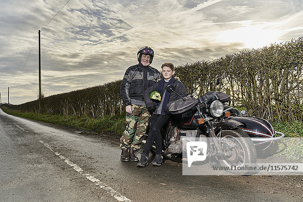 Porträt eines älteren männlichen Motorradfahrers und seines Enkels  die auf einem Motorrad am Straßenrand sitzen