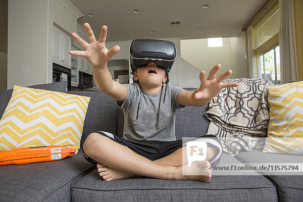 Junge Junge sitzt im Schneidersitz auf dem Sofa  trägt ein Virtual-Reality-Headset  die Hände vor sich ausgebreitet
