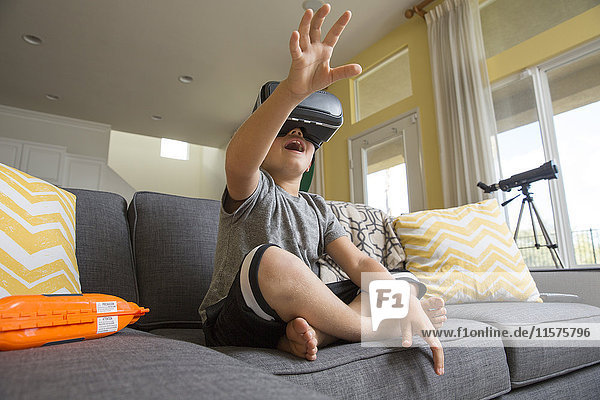 Junge Junge sitzt im Schneidersitz auf dem Sofa  trägt ein Virtual-Reality-Headset  die Hände vor sich ausgebreitet