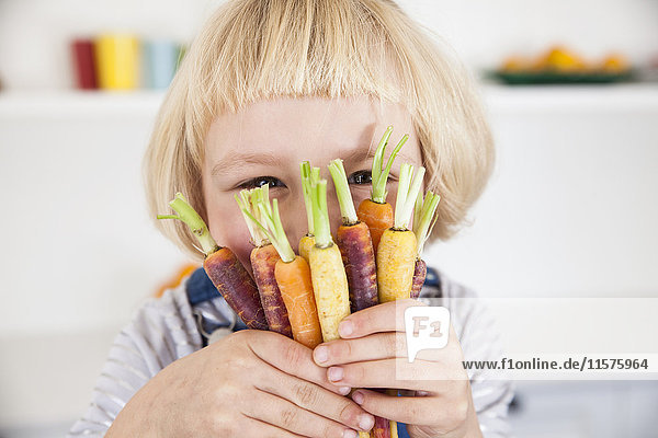 Portrait of cute girl in kitchen holding bunch of colourful carrots to her face