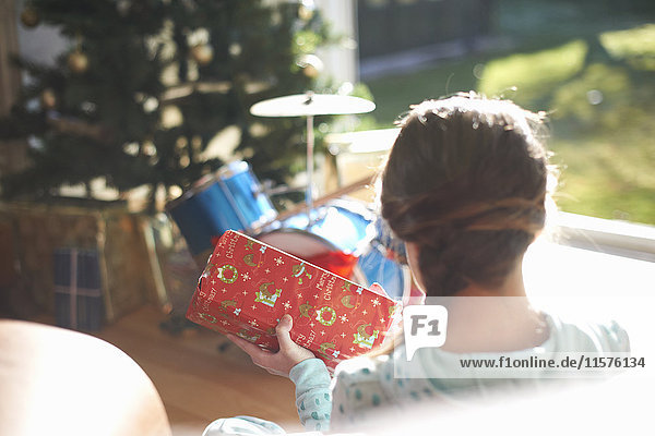 Über-Schulter-Ansicht eines Mädchens  das auf dem Wohnzimmerboden sitzt und ein Weihnachtsgeschenk öffnet