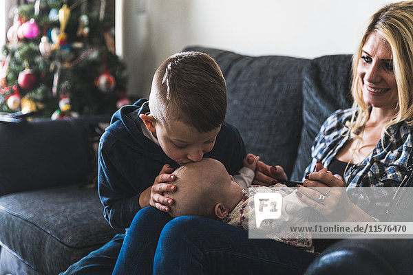 Mother sitting on sofa with young baby on lap son kissing baby on head