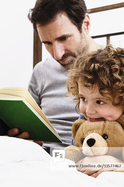 Mädchen umarmt Teddybär  während der Vater im Bett ein Märchenbuch liest