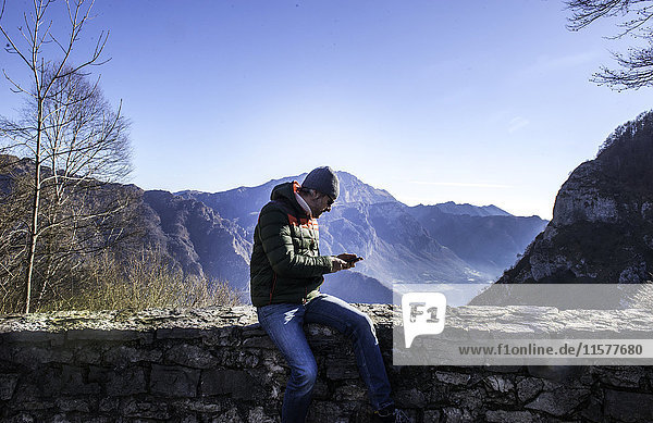 Mann sitzt auf einer Mauer in ländlicher Umgebung und schaut auf ein Smartphone  Italien