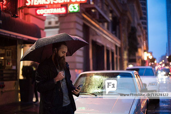Mittelgroßer erwachsener Mann  der nachts in der Stadt spazieren geht  einen Regenschirm benutzt und auf ein Smartphone schaut  Downtown  San Francisco  Kalifornien  USA