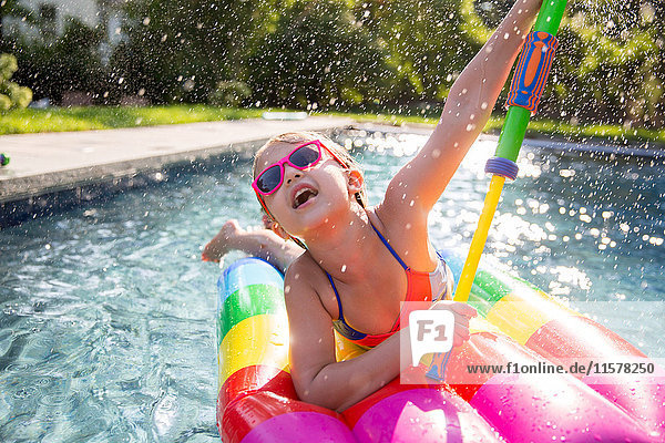Girl in bikini on inflatable playing with water gun in outdoor swimming pool