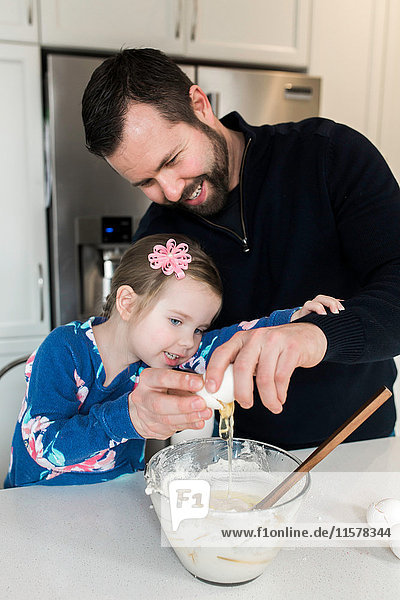 Mittelgroßer erwachsener Mann und Tochter schlagen zusammen ein Ei in eine Rührschüssel an der Küchentheke