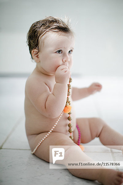Naked female toddler sitting on bathroom floor tasting beads