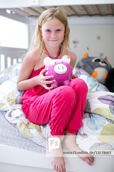 Porträt eines Mädchens  das auf einem Etagenbett sitzt und ein Sparschwein hält