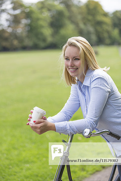 Hübsche blonde Frau mit Fahrrad und Kaffee im Park