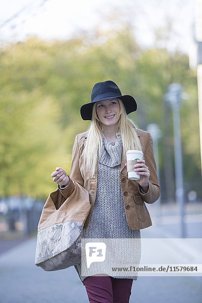 Hübsche blonde Frau mit Hut und Kaffee für einen Spaziergang im Stadtzentrum.