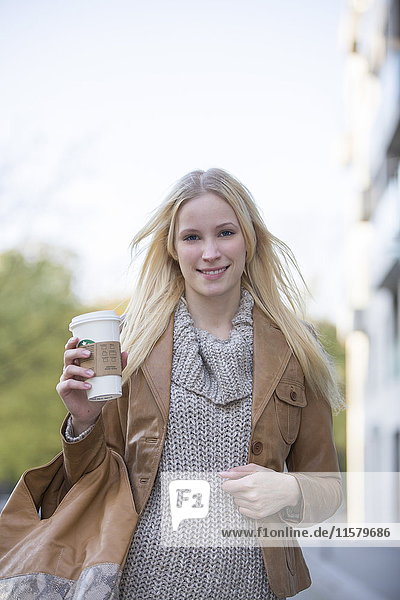 Hübsche blonde Frau mit Café zum Spazierengehen im Stadtzentrum und lächelndem Blick auf die Kamera.