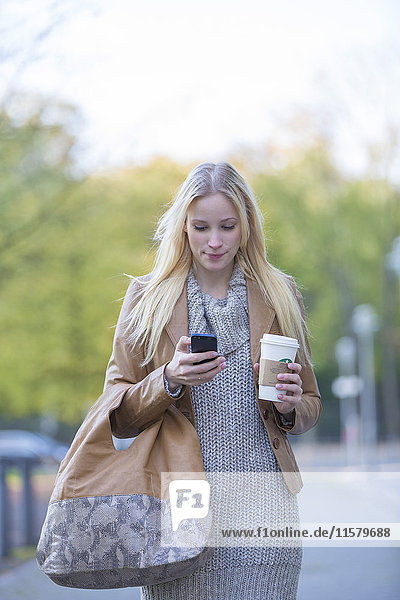 Hübsche blonde Frau  die ihr Handy mit Kaffee überprüft  um im Stadtzentrum spazieren zu gehen.