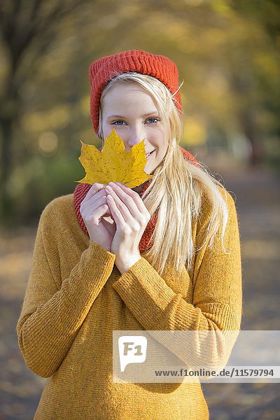 Porträt einer hübschen blonden Frau im Park im Herbst mit Blatt  das teilweise ihr Gesicht bedeckt.