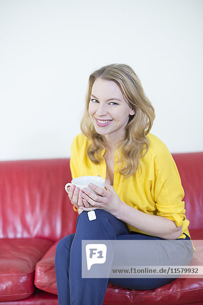 Porträt einer schönen blonden Frau  die eine Tasse Tee auf dem Sofa genießt und vor der Kamera lächelt.