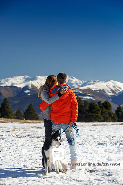 Rückansicht eines romantischen Paares mit Hund beim Blick auf die schneebedeckte Berglandschaft