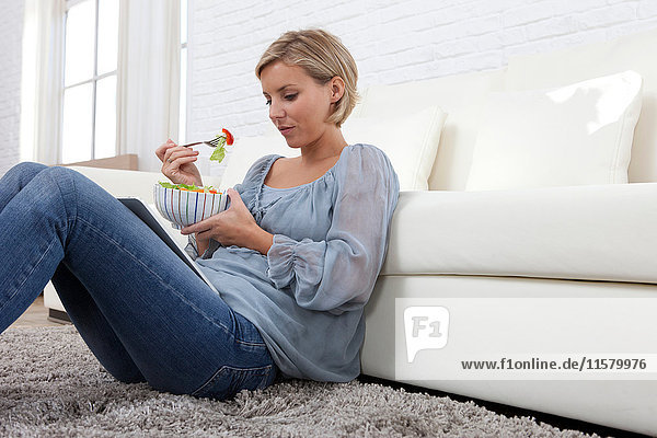 Mittelgroße erwachsene Frau sitzt auf einem Teppich  schaut auf ein digitales Tablett und isst Salat