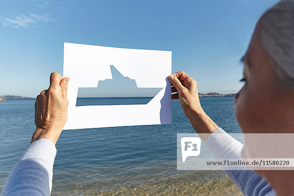 Ältere Frau steht mit dem Gesicht zum Meer und hält ein Papier mit einem Ausschnitt aus einem Boot