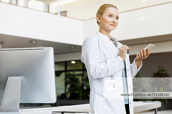 Doctor standing beside computer  holding smartphone