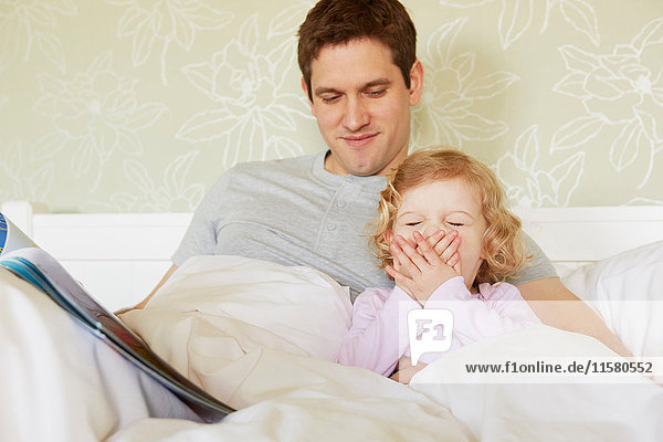 Female toddler yawning in bed with father