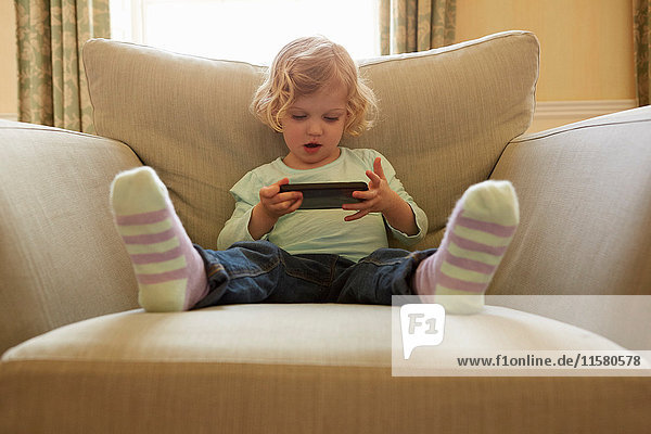 Female toddler sitting on armchair playing hand held computer game