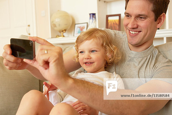 Female toddler sitting on father's knee taking smartphone selfie