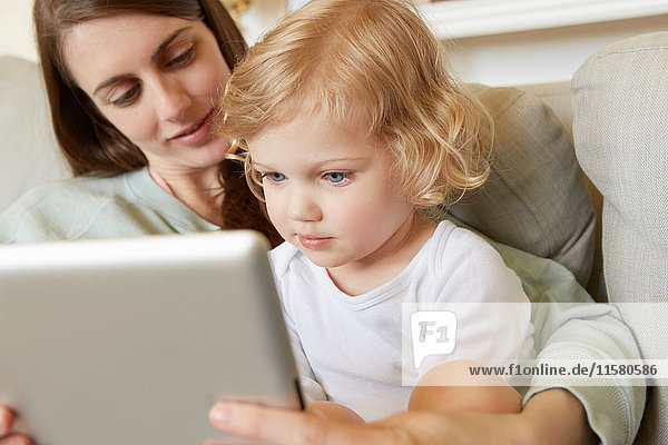 Weibliches Kleinkind sitzt auf dem Knie der Mutter und schaut auf das digitale Tablett